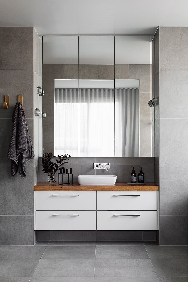 Blick auf Waschtisch mit Tischplatte aus Holz, darüber Spiegelschrank im Bad mit grauen Fliesen