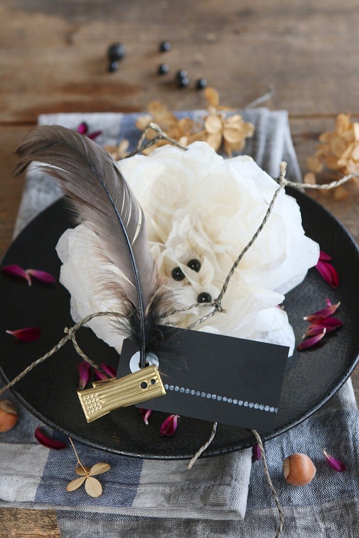 Tischdekoration zu Halloween mit schwarzem Teller, darauf weiße Papierblume, Namensschild mit schwarzer Feder und Goldklammer