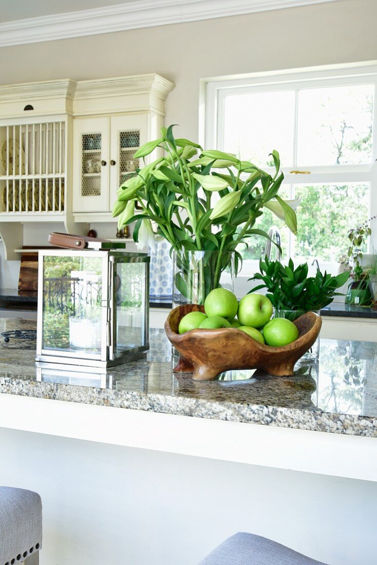 Green apples in fruit bowl, candle lantern and vase of white lilies on kitchen worksurface
