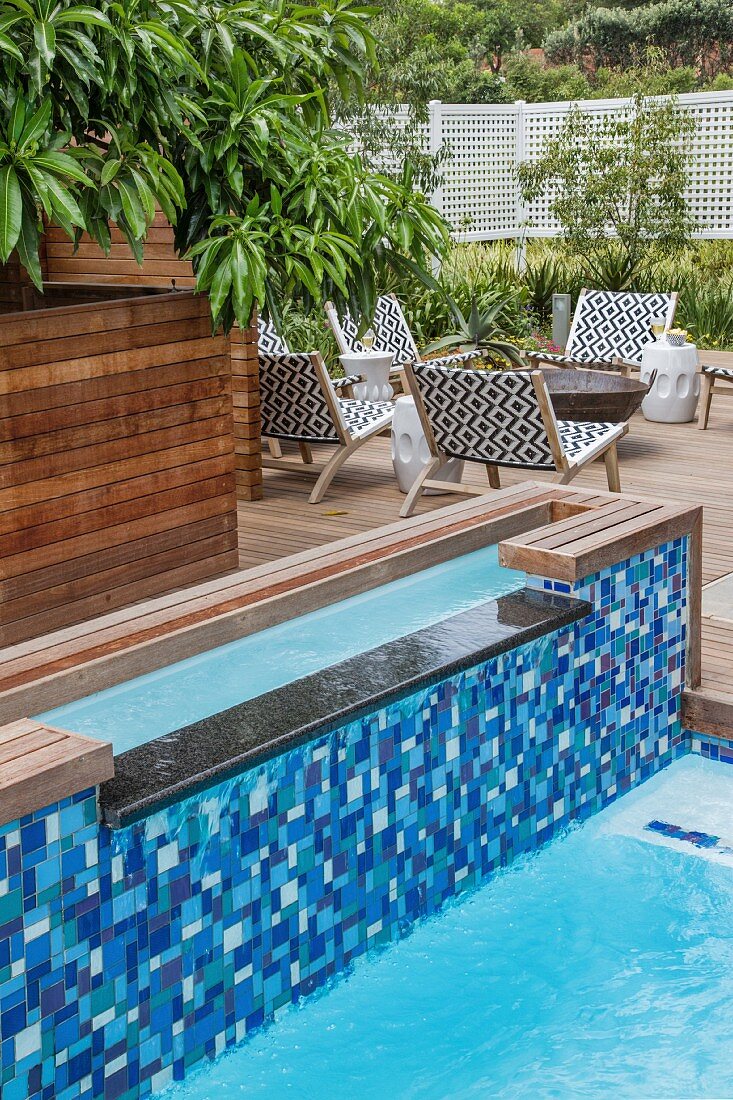 Split pool with mosaic-tiled wall in shades of blue; outdoor chairs around fire pit in background