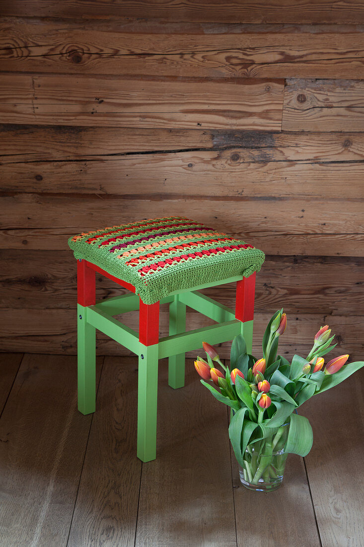 Crocheted seat cover on red and green stool against wooden wall