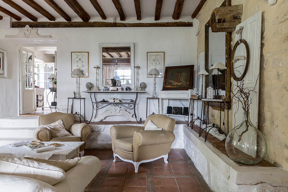 Beigefarbene Sessel und Couch in rustikalem Wohnraum mit gemauerter Ablagefläche, Terrakottafliesenboden und Holzbalkendecke