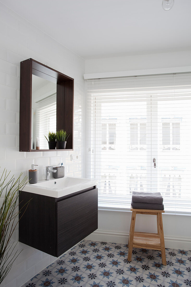 Sink and mirror on white tiled wall, wooden stool in front of window with louvre blinds in bathroom