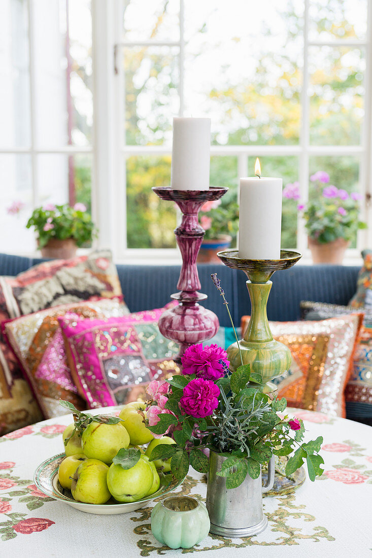 Runder Tisch mit Obst, Blumen und Kerzen, dahinter Sofa mit bunten Kissen im Wintergarten