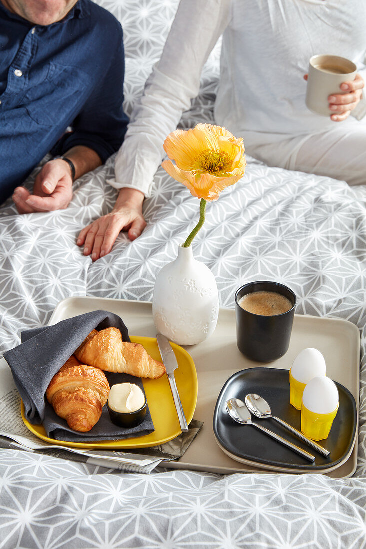 Frühstückstablett mit Croissant, Eiern, Kaffee und gelber Mohnblüte auf dem Bett, Pärchen im Hintergrund