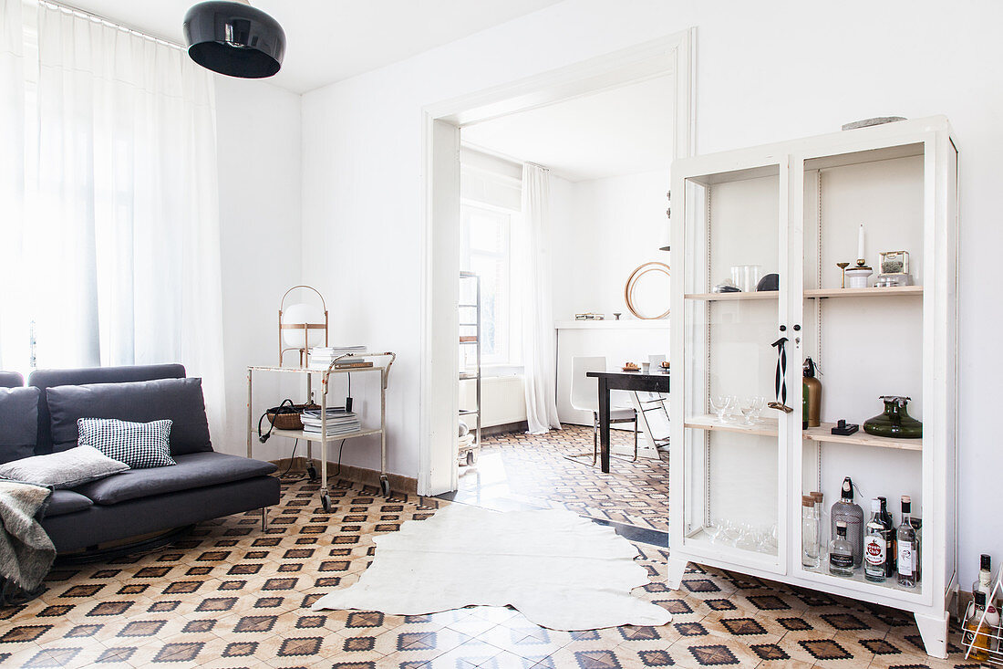 Cowhide rug on patterned tiled floor, display cabinet, grey sofa and vintage serving trolley in living room with open doorway