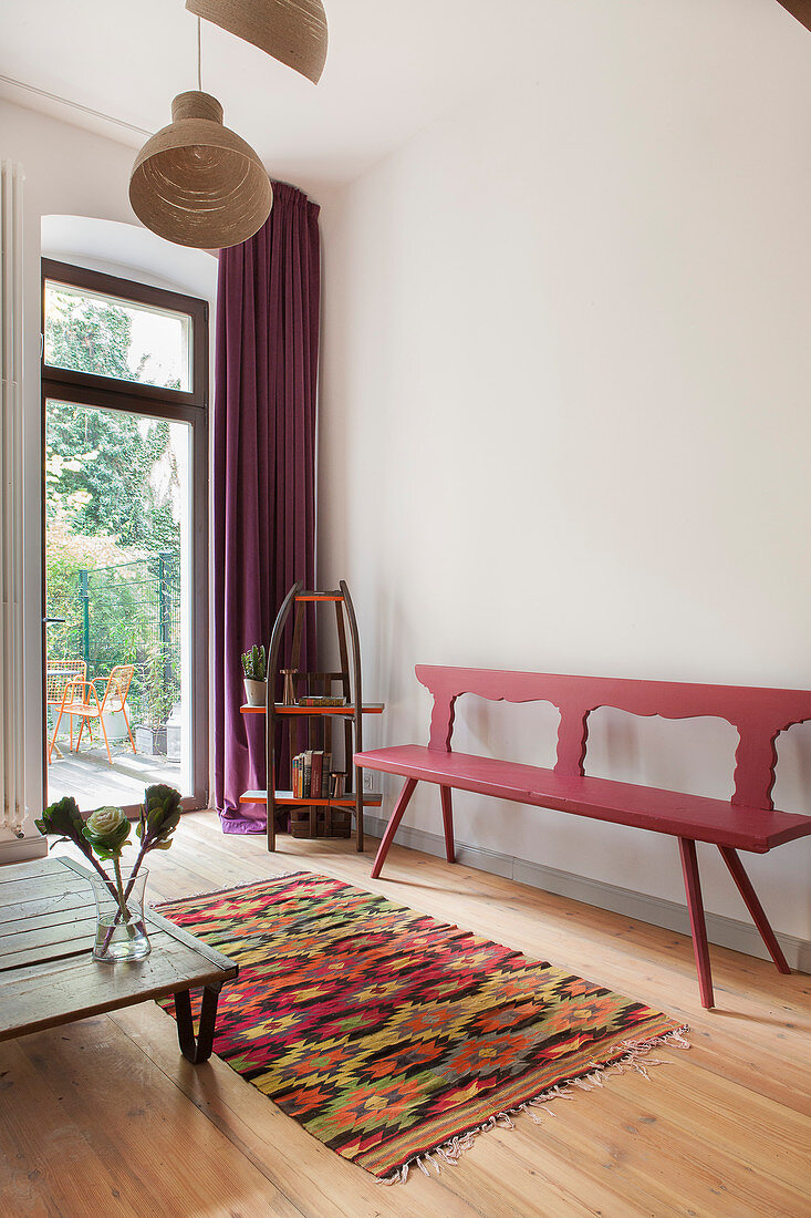 Rot lackierter Holzbank, Vintage Bücherregal, niedriger Couchtisch und bunter Teppich vor Terrassentür im Wohnzimmer
