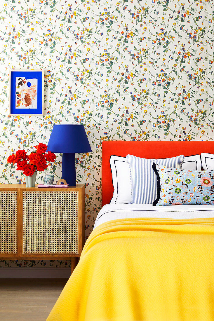 Bett mit orangefarbenem Betthaupt und gelber Tagesdecke, daneben Nachtschränckchen im Schlafzimmer mit Blumentapete