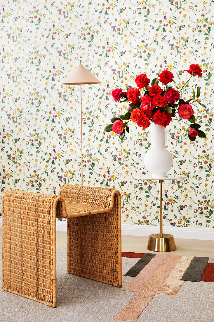 Rattan table, floor lamp and side table with a bouquet of red roses in front of floral wallpaper