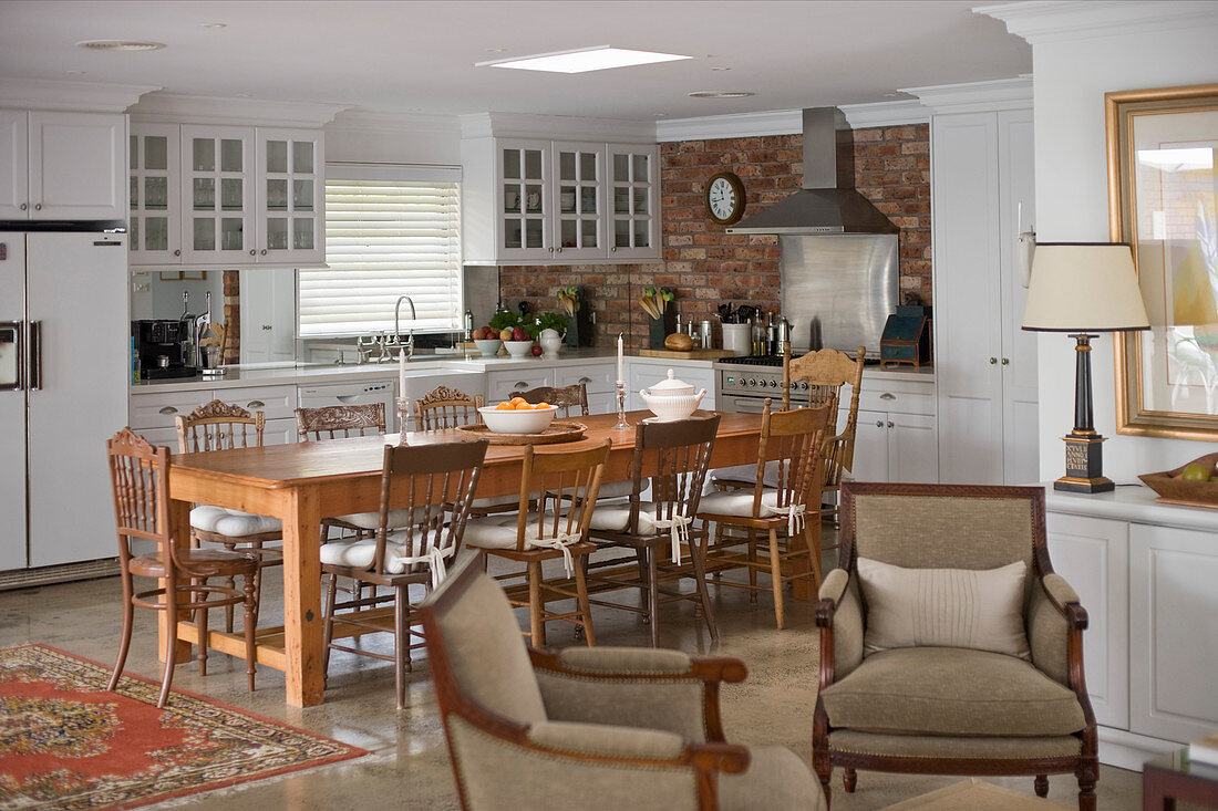 Kitchen with white cabinets and brick wall behind solid wooden dining table with chairs and two armchairs