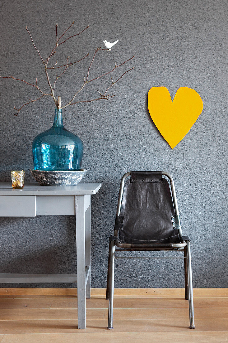 Vintage Stuhl, türkisfarbene Ballonflasche mit Zweig auf Schreibtisch und gelbes Herz an grauer Wand