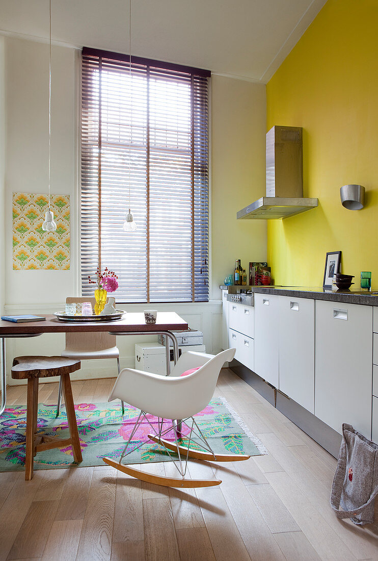 Klassiker Schaukelstuhl, Tisch und weiße Unterschränke in Küche mit gelber Wand  mit Jalousie am Fenster