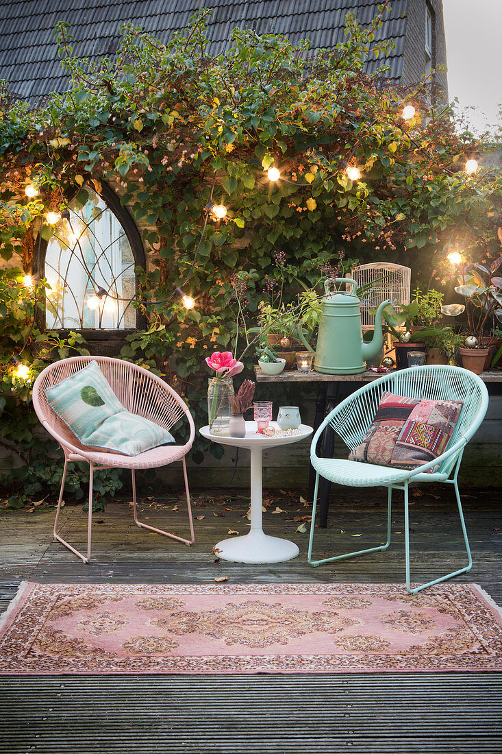 Retro easy chairs and side table on terrace surrounded by plants and fairy lights