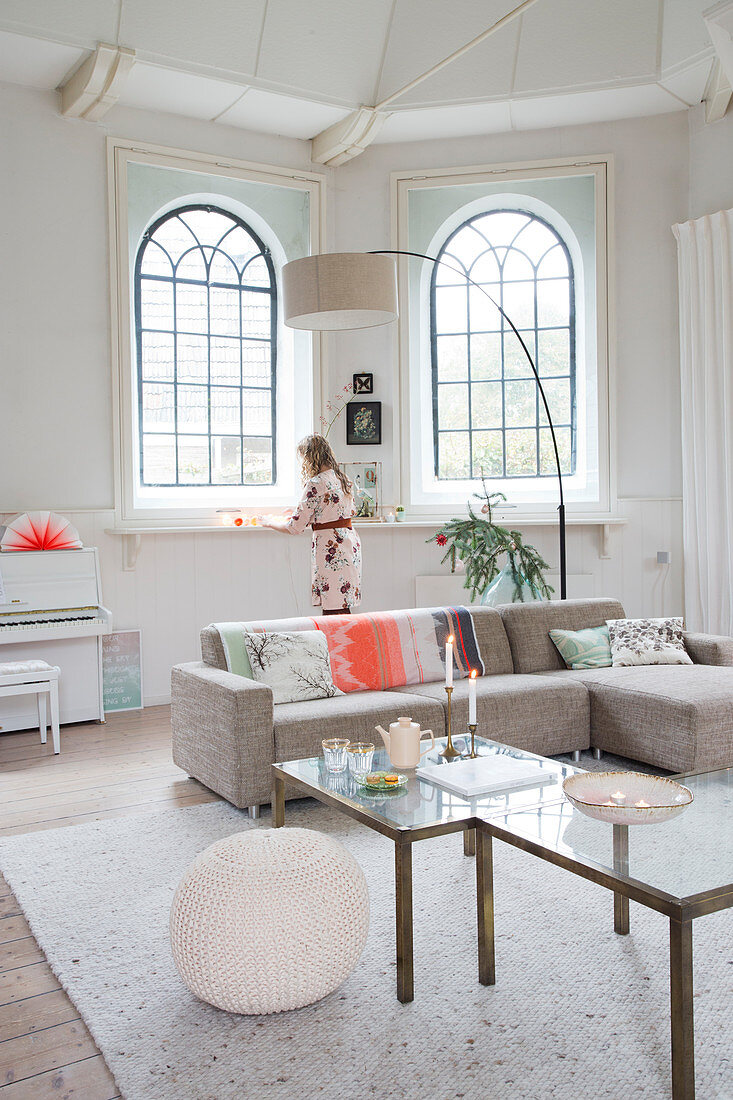 Sofa, set of coffee tables and arc lamp in front of arched windows in living room with woman in background