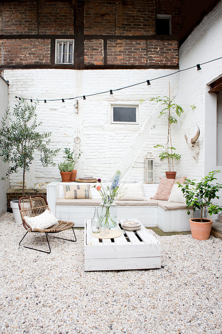 Holzbank, Palettentisch und Stuhl auf Terrasse mit weiß gestrichener Ziegelwand und Kiesboden