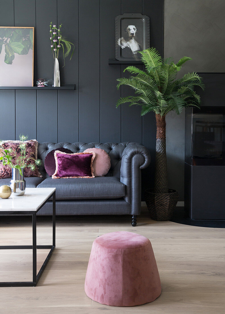 Dunkles Sofa mit Rosa Kissen vor dunkler Wand, Sitzpouf im Vordergrund