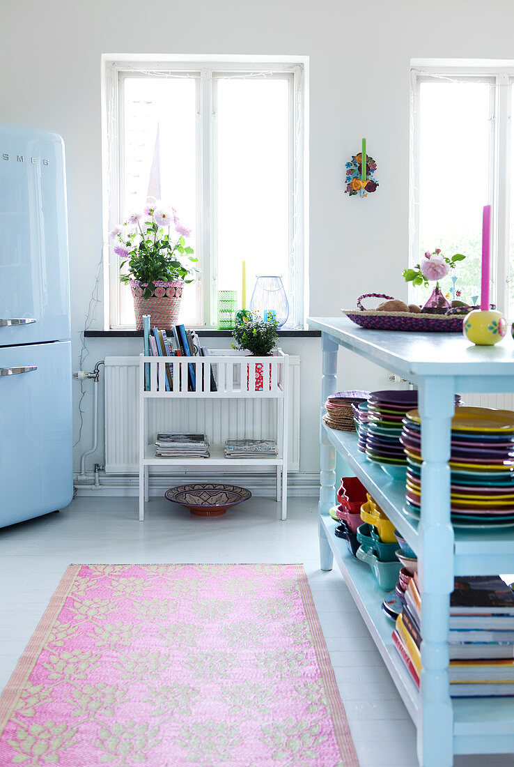 Hellblaues, offenes Regal mit buntem Geschirr in Wohnküche, im Hintergrund Kühlschrank neben Fenster