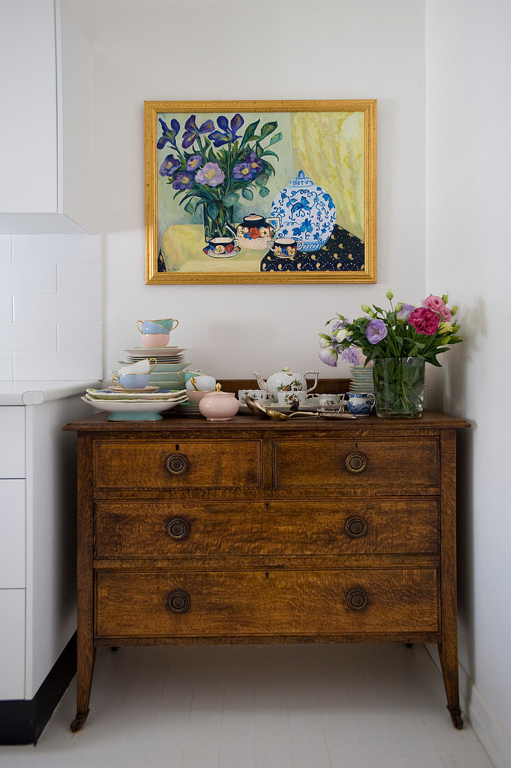 Crockery and vase of flowers on rustic chest of drawers below painting