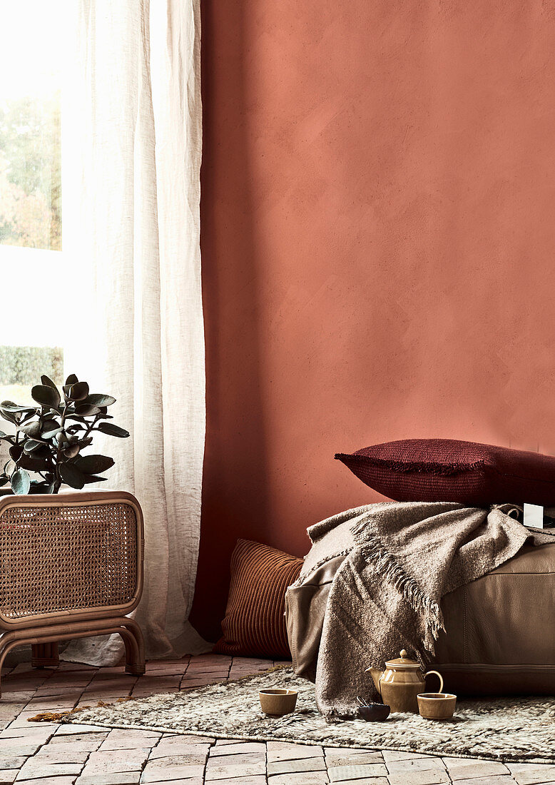 Kissen, Decke und Teegeschirr auf Teppichvorleger vor ziegelroter Wand, Zimmerpflanze vor Fenster