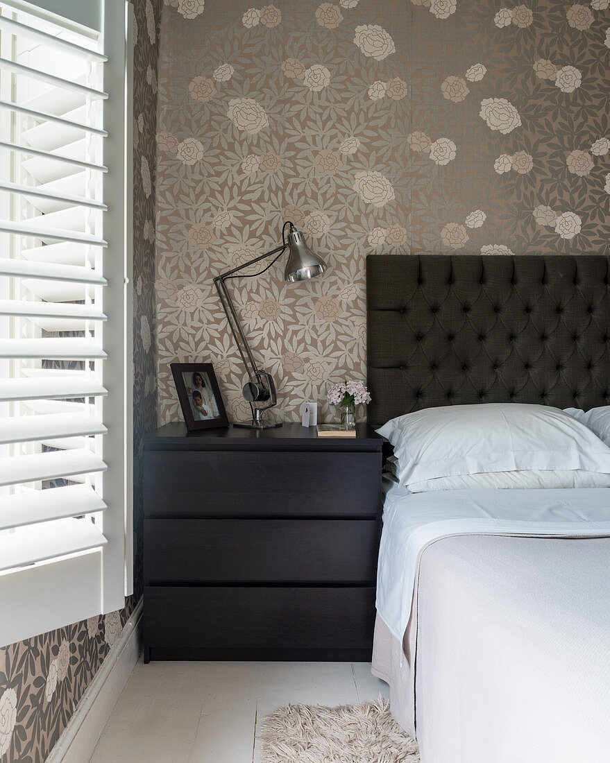 Bed with black headboard and black bedside cabinet against beige floral wallpaper