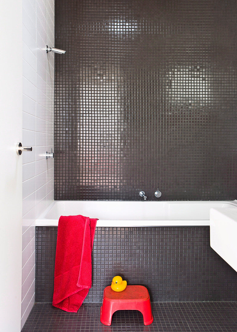 Badezimmer mit grauen Mosaikfliesen, roter Hocker vor Badewanne