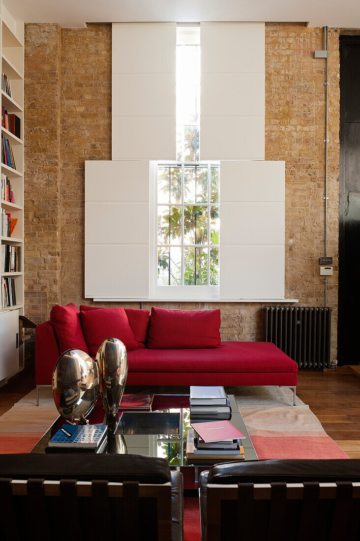 Red chaise longue below floor-to-ceiling windows with sliding shutters in brick wall