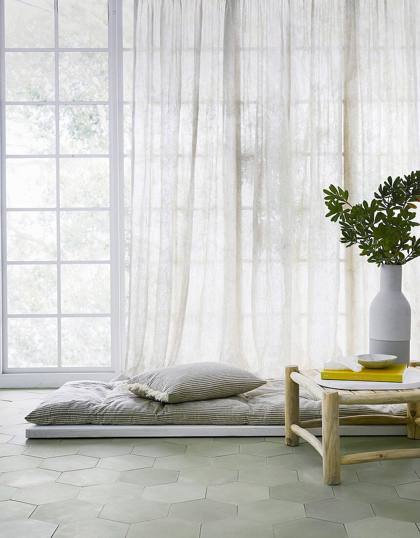Bodenmatte mit Kissen vor Fensterfront daneben Beistelltischchen mit Vase