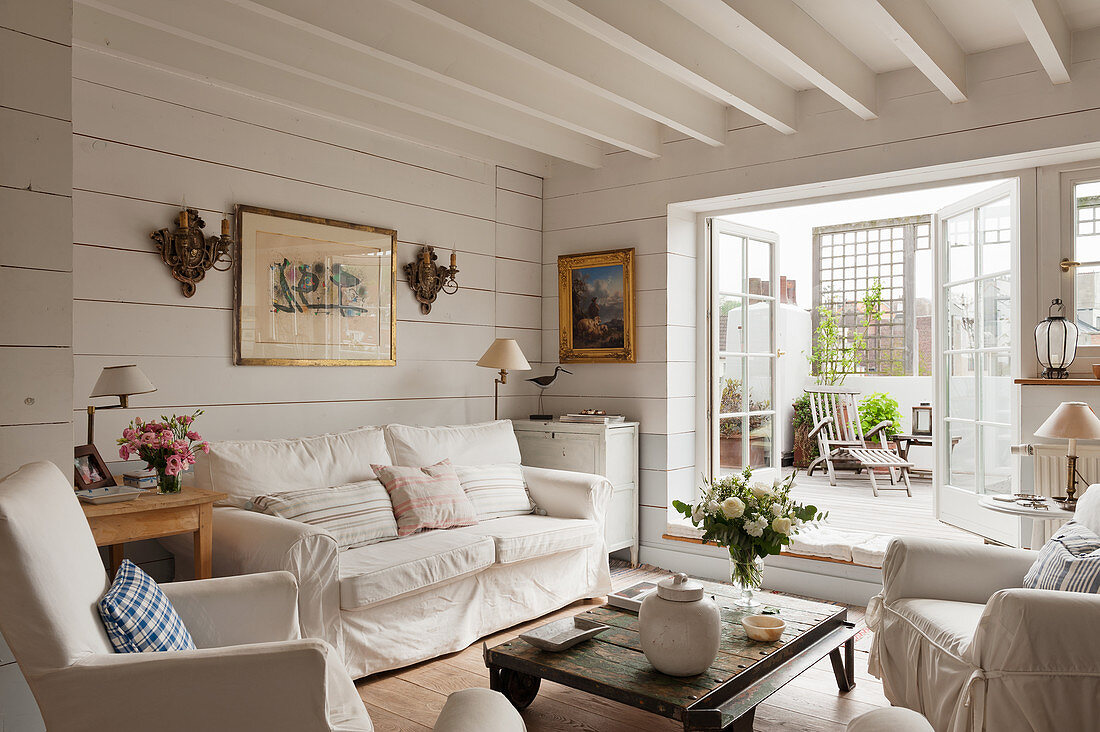Weiße Hussenmöbel und Couchtisch auf Rädern im Wohnzimmer mit weiß gestrichenen Holzwänden und Terrassenzugang
