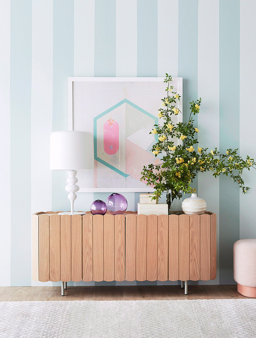 Sideboard mit Tischleuchte, Glasdeko und Blütenzweig vor gestreifter Wand