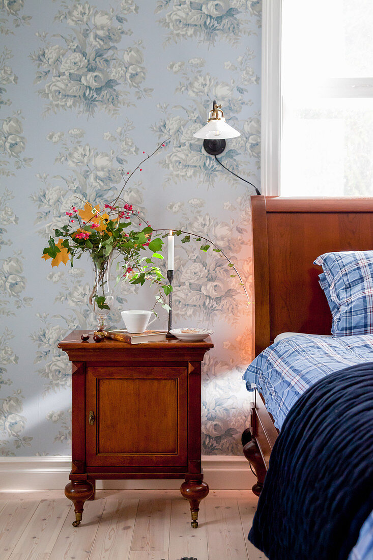 Antique bedside cabinet next to wooden bed in bedroom with vintage-style floral wallpaper