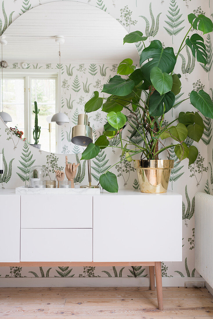 Weißes Sideboard mit großer Zimmerpflanze, darüber runder Spiegel, Tapete mit botanischem Muster