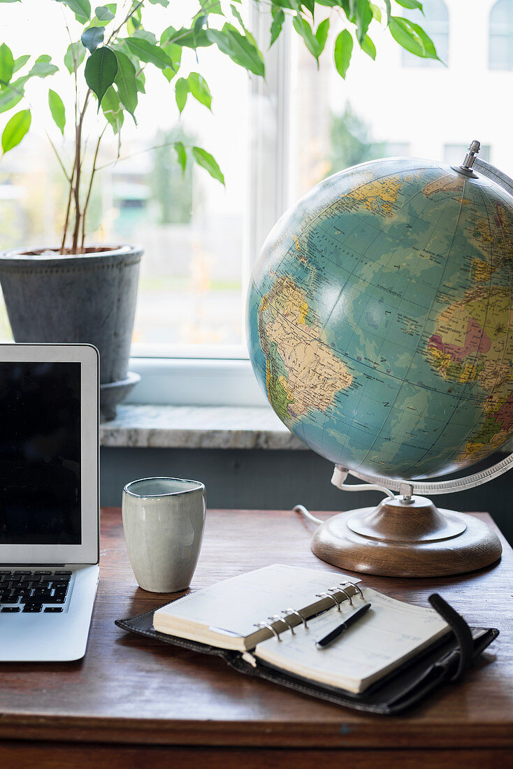 Globus neben Laptop und Taschenkalender auf Schreibtisch am Fenster