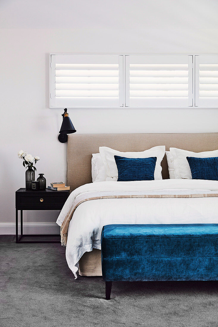 Blaue Polsterbank vor dem Bett unter horizontalen Lamellenfenstern