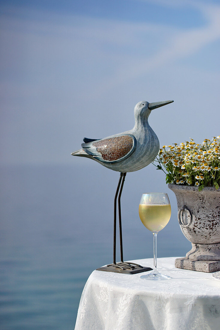 Vogelfigur und ein Glas Weißwein auf Tisch, im Hintergrund das Meer