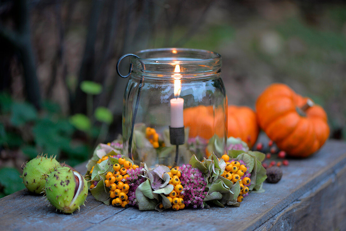 Windlicht mit Kränzchen aus Fetthenne, Hortensienblüten und Vogelbeeren, daneben Zierkürbisse und Kastanien