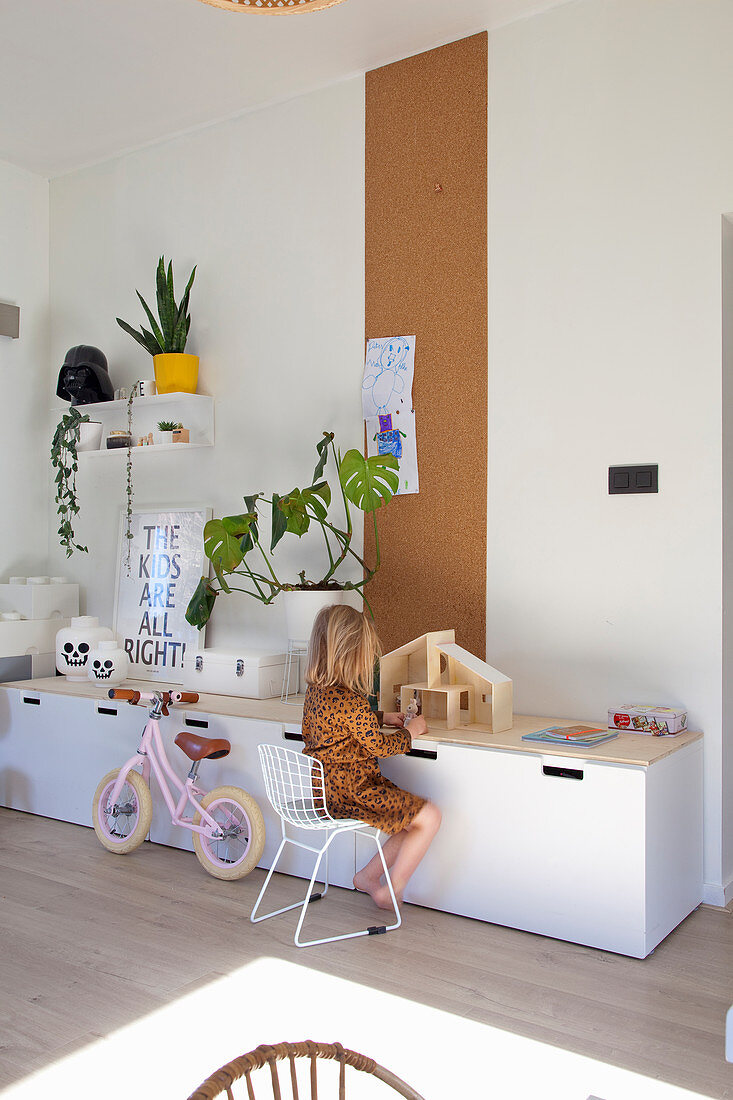 Child sitting at sideboard playing with dolls' house in front of strip of cork pinboard on wall