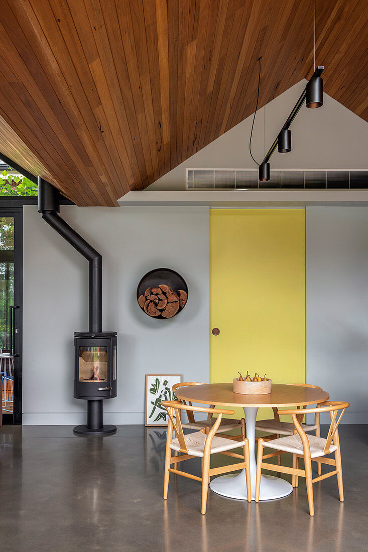 Living room under a pointed wooden roof with a round dining table and cast iron wood stove
