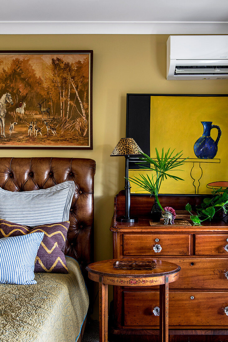 Hunting paintings over a leather bed next to a chest of drawers and wooden side tables