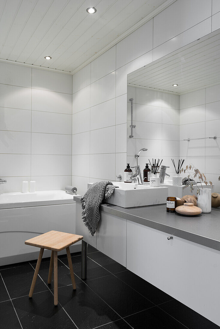 Sink below large mirror in bathroom with black floor tiles and walls entirely tiled in white