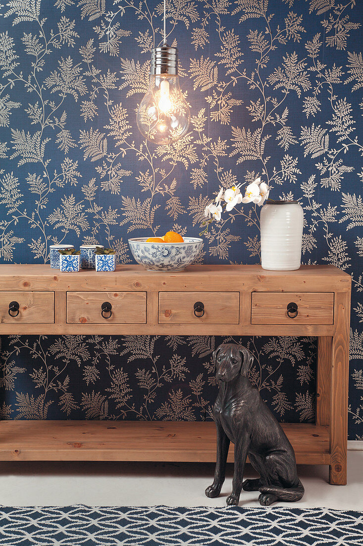 Holzkonsole mit Schubladen, Glühbirne und Hundeskulptur vor blauer Tapete mit Blättermotiv