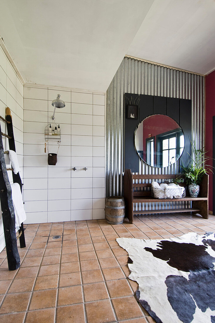 Holzbank und runder Spiegel vor Wellblechwand, Tierfellteppich auf Fliesenboden in großzügigem Badezimmer