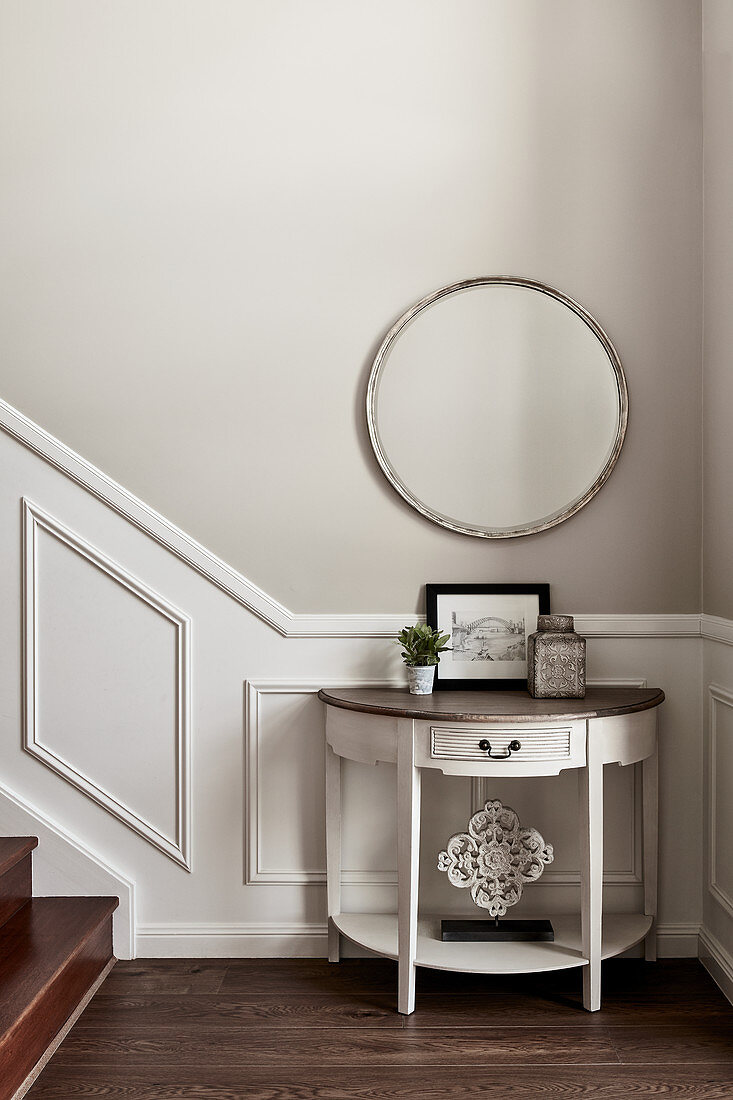Semicircular console table against panelled wainscoting below round mirror at foot of staircase