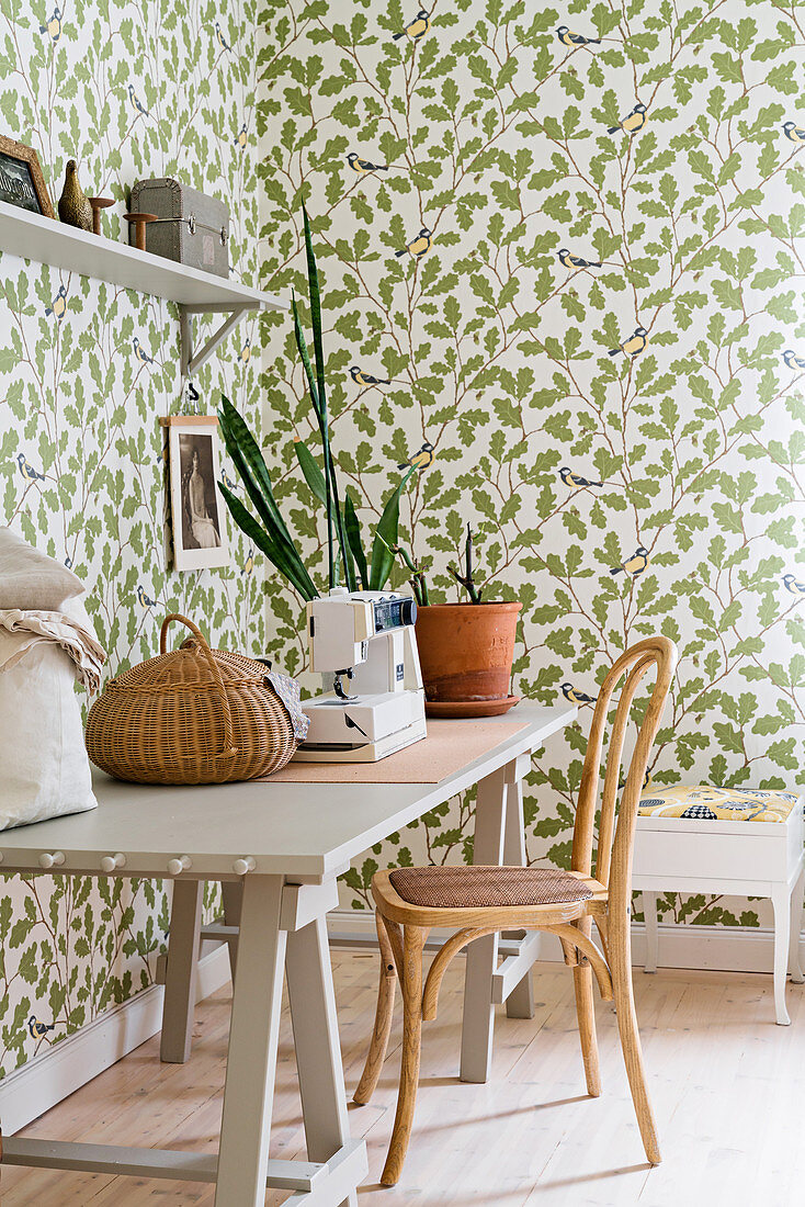 Sewing machine on wooden table, chair and leaf-patterned wallpaper in bright room