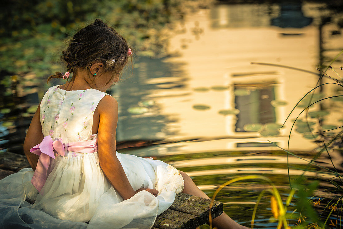 Mädchen im weißen Sommerkleid sitz auf Steg an idyllischem Teich