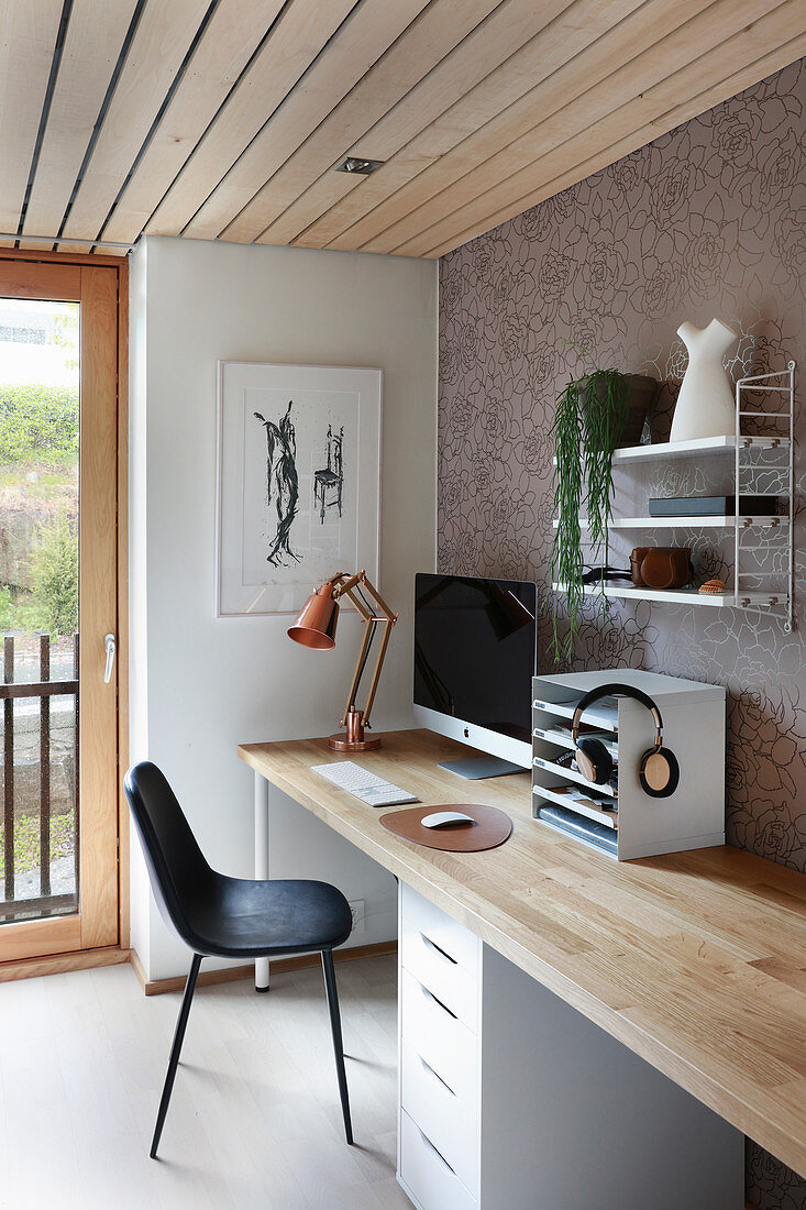 Schreibtisch mit heller Holzplatte, darüber Regal an tapezierter Wand