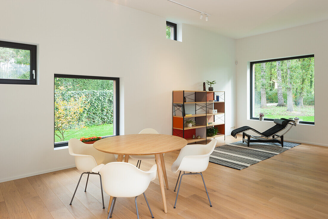 Dining room with round wooden table and white chairs, lounger in front of window in the background