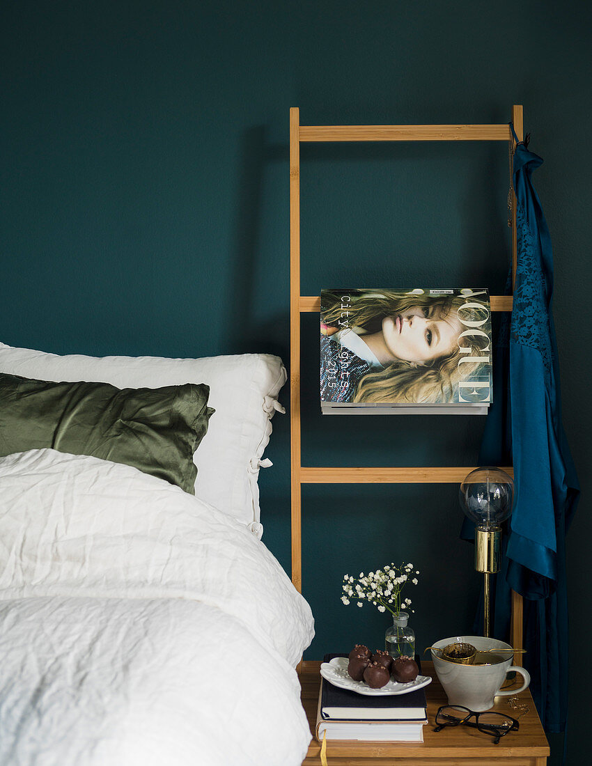 Weiß bezogenes Bett vor taubenblauer Wand mit Holzleiter und Modezeitschrift