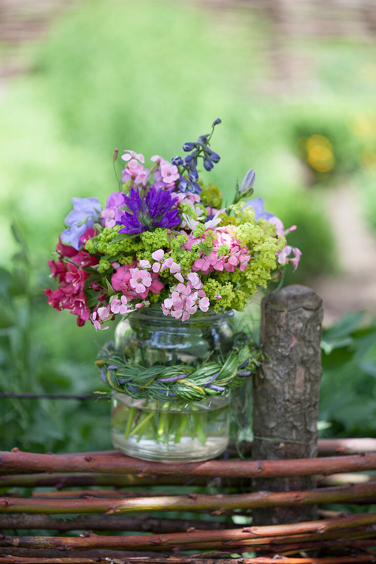 Blumenstrauß im Einmachglas mit Zopf aus Gras auf Flechtzaun gestellt