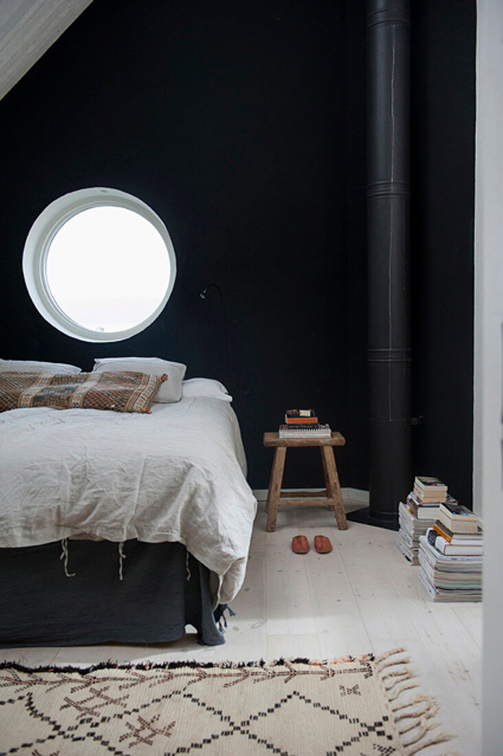 Bed below round window in black bedroom wall