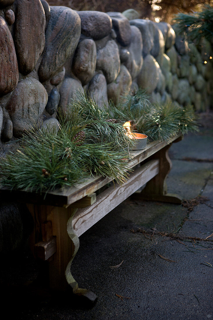 Nadelzweige und Windlicht als winterliche Dekoration auf Holzbank vor Natursteinmauer